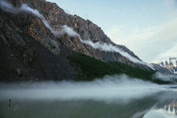 Scenic alpine landscape with long low cloud on high rockies in sunlight above green mountain lake in fog. Atmospheric mountain scenery with long low clouds on green water of mountain lake and rocks.