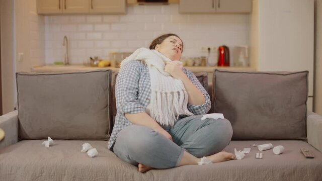 Sick overweight woman in scarf coughing, sitting on sofa at home, seasonal virus