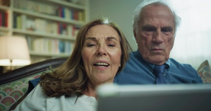 Cinematic shot of happy modern mature senior couple is using a tablet for family entertainment while sitting on a sofa in living room at home.Concept:technology, modern generation, connection, retired