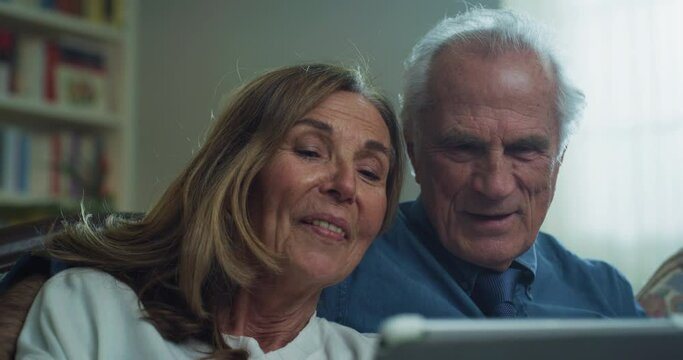 Cinematic shot of happy modern mature senior couple is using a tablet for family entertainment while sitting on a sofa in living room at home.Concept:technology, modern generation, connection, retired