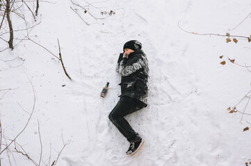 Fototapeta na wymiar A very drunk homeless man in a black coat lies sleeping on his side on white snow in a cold frosty winter with a bottle of alcohol. Photography, concept, copy space.