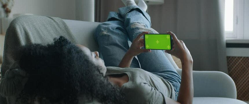 GREEN SCREEN CHROMA KEY DOLLY IN Black African American female lying on sofa, holding phone in hands. Horizontal orientation. Touch clicking center of the screen