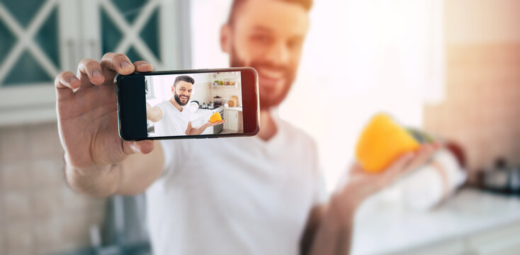 Young handsome happy bearded man with smart phone in the kitchen at home while he is preparing the healthy fresh vegan salad and making selfie photo