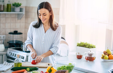 Happy smiling cute woman is preparing a fresh healthy vegan salad with many vegetables in the...