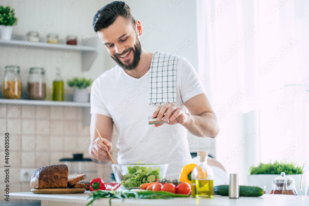 Wall mural handsome happy bearded man is preparing wonderful fresh vegan salad in the kitchen at home