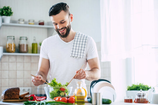 Handsome happy bearded man is preparing wonderful fresh vegan salad in the kitchen at home