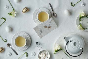 Springtime afternoon tea. Spring freesia flowers, white teapot and tea cup on table. Flat lay, off white textile tablecloth. Easter eggs, white ceramic cups and sugar hearts.