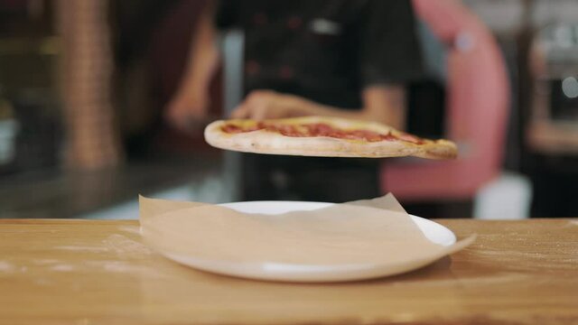Delicious ready pizza lying on round plate on wooden table. Blur background of pizzaiolo in black uniform with long shovel in hands. Cooking process at restaurant.
