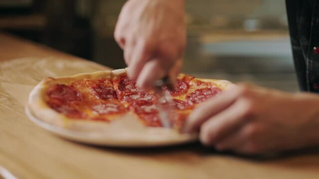Close up of male hands cutting freshly prepared pizza with special knife on restaurant kitchen. Chef slicing tasty pizza on wooden table. Italian culinary.