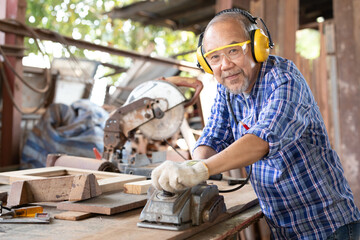 portrait senior asian man carpenter wearing glasses and headphone, using electric wood planers on a piece of wood in workshop - obrazy, fototapety, plakaty