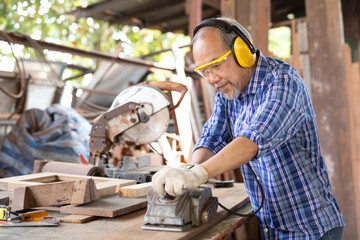 senior asian man carpenter wearing glasses and headphone, using electric wood planers on a piece of wood in workshop