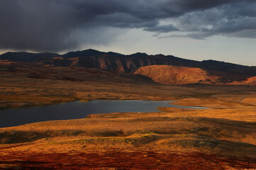 Colorful sunset above highland steppe shore of lake with dry yellow grass on the background of rocky mountains under storm sky