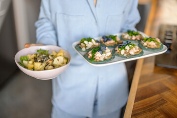 Woman in blue shirt hold a set of appetizer on a plate with seaweed, cheese, blueberry and hummus and freshly cooked salad on kitchen background. Homemade tasty snack.
