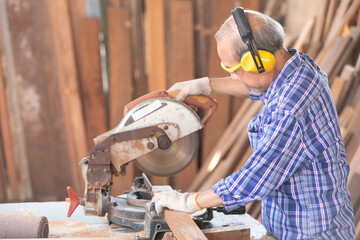 senior asian man carpenter wearing glasses and headphone, using electric circular saw for cutting...