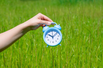 Young female hand holds sunlit alarm clock on green blurred nackground with bokeh effect. Concept and metaphor of summer time coming.