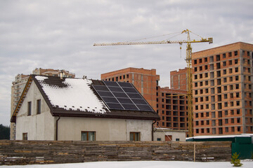 small concrete house on the background of a large construction site. solar panels on the roof. tower crane construction site