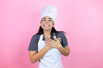 Young hispanic woman wearing baker uniform over pink background smiling with her hands on her chest and grateful gesture on her face.
