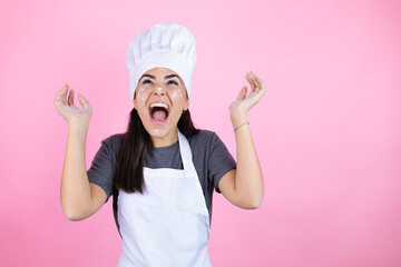 Young hispanic woman wearing baker uniform with flour on the face over pink background celebrating crazy and amazed for success with arms raised and open eyes screaming excited.winner concept