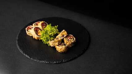 Custom sushi roll in tempura with nori, fresh salmon, tuna, avocado, masago caviar, drizzled with pineapple sauce with salad pouring as decoration on a black plate on a black table and background.