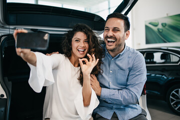 Happy middle age couple enjoying while choosing and buying new car at showroom. They making selfie photo with their new car.