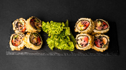 Custom sushi roll in tempura with nori, fresh salmon, tuna, avocado, masago caviar, drizzled with pineapple sauce with salad pouring as decoration on a black plate on a black table and background.