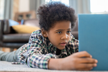 Cute elementary schoolboy with digital tablet lying on the floor against couch