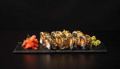Custom sushi roll with nori, cuttlefish ink, fresh salmon, cream cheese, pepper pumpkin, eel, unagi sauce and edible gold leaf with red ginger, wasabi on a square plate on black table and background.