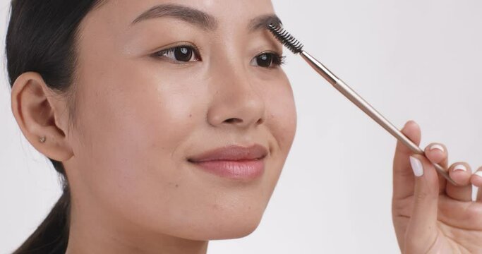 Young asian lady styling her eyebrows with eyebrow mascara, white studio background, close up, slow motion
