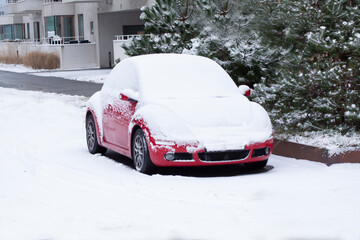 Red car covered with snow with pine trees and an apartment building in background.