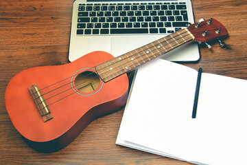 Hawaiian four-string guitar Ukulele on wooden table, laptop, notepad and pen. Concept of learning to play musical instrument, online guitar courses.