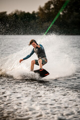 young sportsman in vest stands on wakeboard and ride down on the splashing wave.