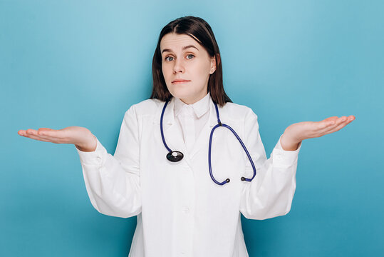 Portrait of indecisive woman doctor in white coat, shrugging and looking troubled answer, dont know, isolated on blue wall. Covid-19, coronavirus outbreak, healthcare workers and pandemic concept