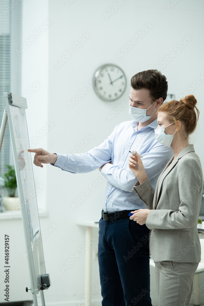 Poster young manager in protective mask pointing at whiteboard during presentation