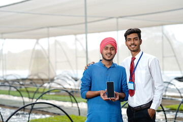 Young indian farmer with agronomist showing smartphone at poly house or greenhouse