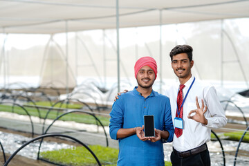 Young indian farmer with agronomist showing smartphone at poly house or greenhouse