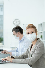 Young female office manager in protective mask and elegant suit using laptop