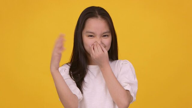 Studio portrait of young frowning girl closing her nose and waving hand, feeling terrible scent, orange background