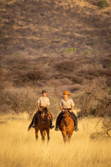 Two women ride through grassland near hill