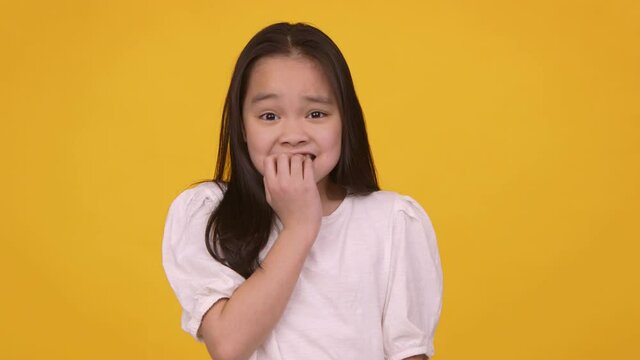 Little asian girl feeling panic, biting her nails nervously, orange studio background, slow motion