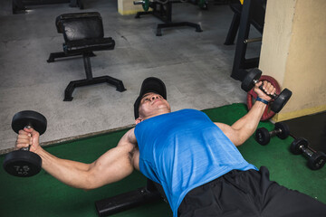 An asian man in a blue tank top does a set of dumbbell chest flys on a flat bench. Training pecs...