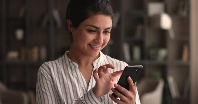 Smiling young indian woman enjoying using mobile software applications, communicating distantly in social networks, shopping in internet store, playing games on cellphone or web surfing information.