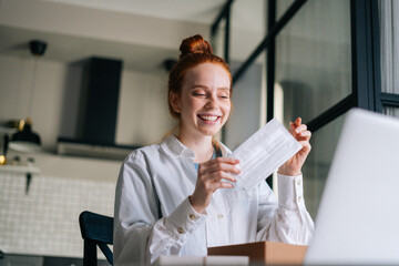 Low-angle view of cheerful redhead young woman wearing medical protective face mask during video call via laptop webcam. Concept of leisure activity red-haired female at home.