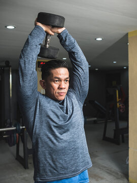 A Fit Athletic Asian Guy In A Sweatshirt Does Standing Overhead Dumbbell Tricep Extension With Both Arms. Starting Position. Working Out And Training Triceps. Open Air Gym Setting.