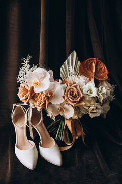 Wedding. The bride's bouquet of brown flowers with a ribbon stands on a dark background next to high-heeled shoes