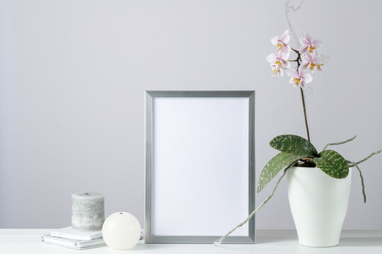 Poster mockup or silver photo frame in modern scandinavian style with pink orchid flowers in the ceramic pot on white wooden table. Home interior decoration
