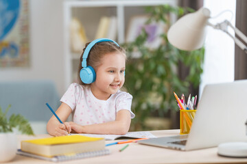 smart smiling little girl wearing headphones is learning online, using laptop remote learning. child listens to teacher and writes down task in notebook. interactive E-Education Distance Home learning