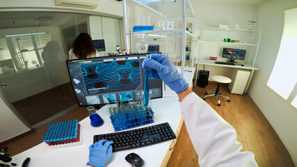 FPV of biologist doctor checking liquide from test tubes, writing on pc, while colleague bringing blood samples. Scientists examining vaccine evolution in medical lab using high tech for research.