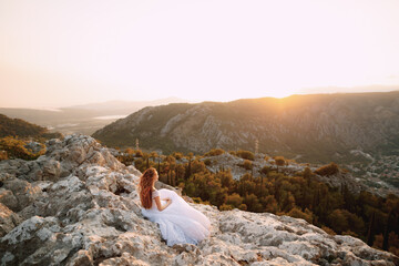The bride in tender wedding dress sits on the rock on Mount Lovcen and looks at the Bay of Kotor 