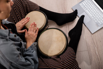 A faceless woman sits on the floor at home and watches educational videos of playing mini bongs. Girl remotely learns to play traditional ethnic drums.
