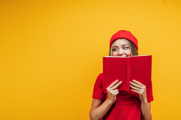 Portrait of a female student in red clothes on a yellow background, she covers her face with a black book and looks at the place for text. Copy space. Background